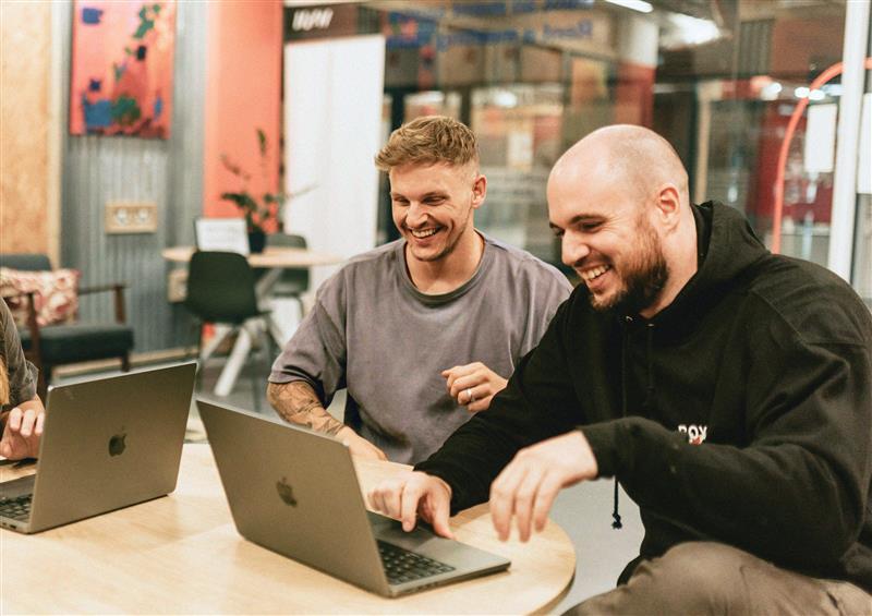 Två män ler och arbetar på bärbara datorer tillsammans vid ett runt bord i en avslappnad YH Akademin kontorsmiljö.