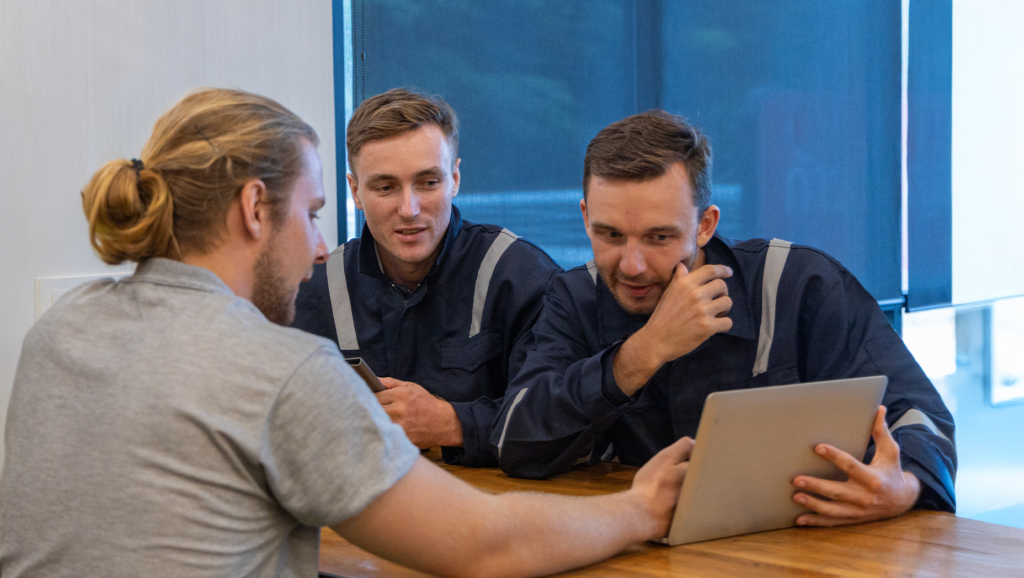 Vid ett bord sitter tre män, varav två i uniform, och tittar på en surfplatta tillsammans och diskuterar något.