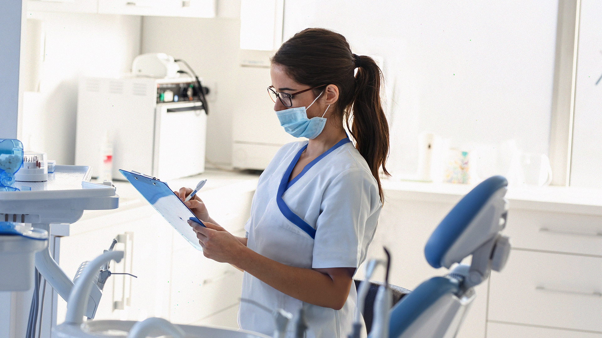En kvinnlig tandläkare iförd mask skriver på en skrivplatta i en ljus tandläkarmottagning.
