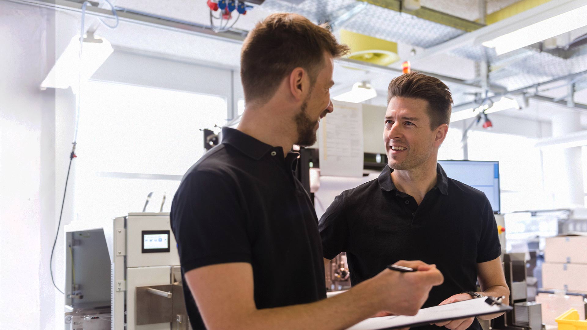Två män i svarta skjortor pratar och ler i ett ljust, modernt kontor eller labb, den ena håller i en skrivplatta.