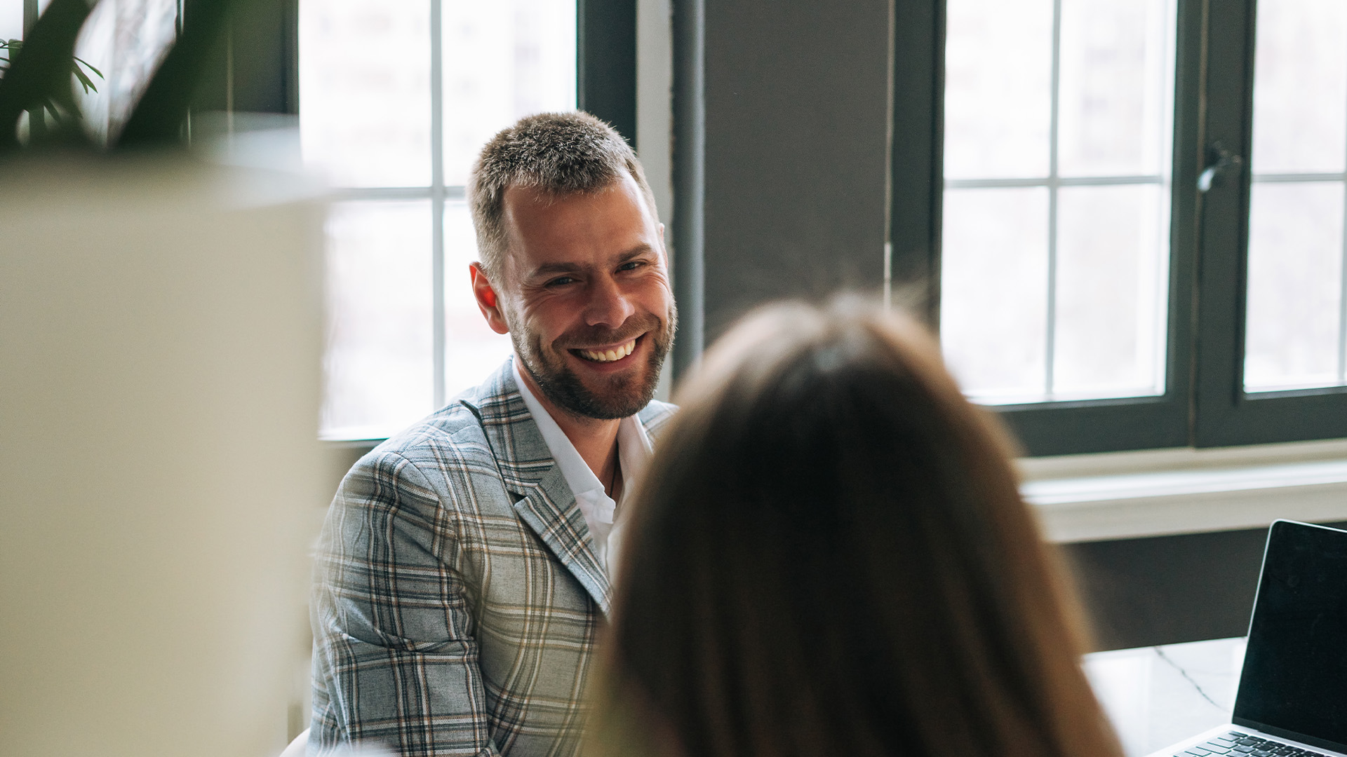 En man i rutig kavaj ler medan han pratar med en kvinna på ett kontor med stora fönster.