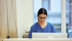 En vårdpersonal i blå operationskläder sitter vid ett skrivbord och arbetar på en bärbar dator i ett ljust kontor.