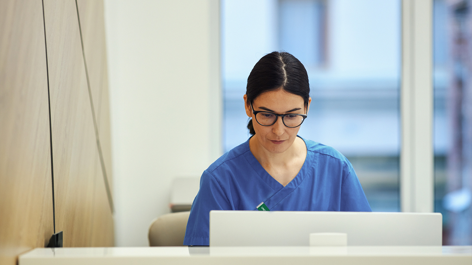 En vårdpersonal i blå operationskläder sitter vid ett skrivbord och arbetar på en bärbar dator i ett ljust kontor.