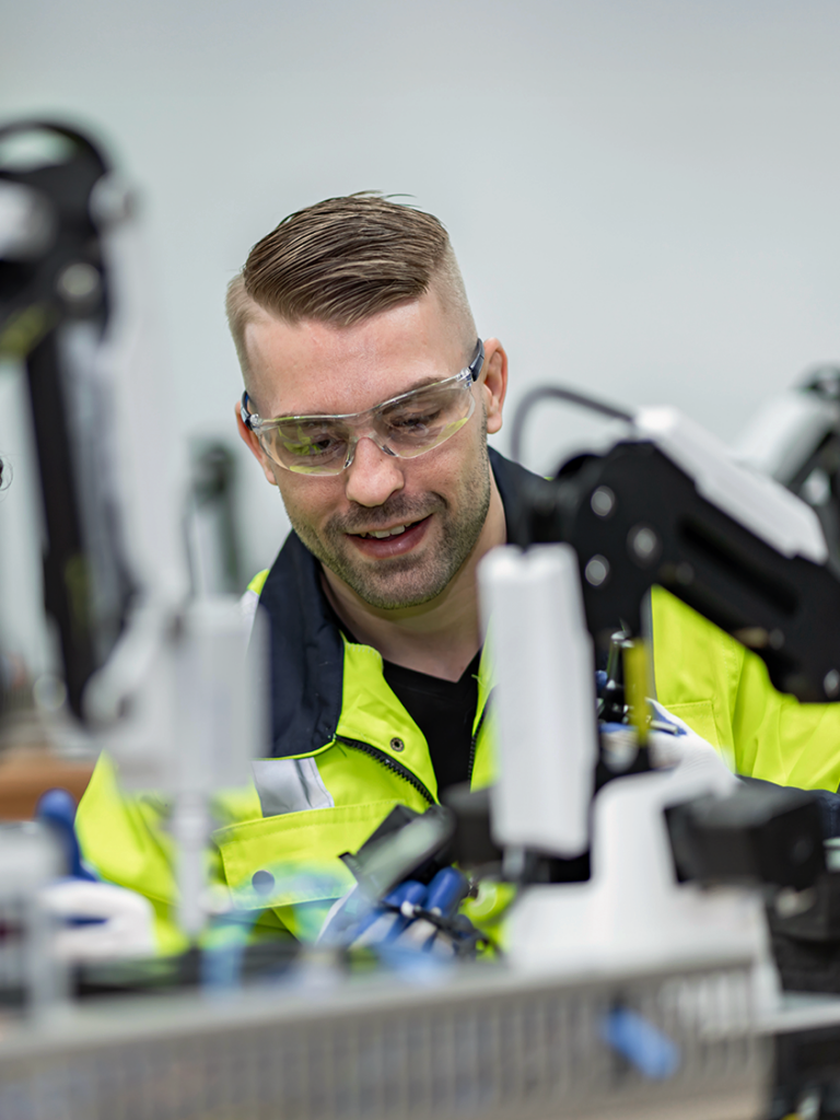 En man i skyddsglasögon och gul jacka manövrerar industrirobotutrustning i ett labb, vilket visar den praktiska utbildning som är typisk för en yrkeshögskola.