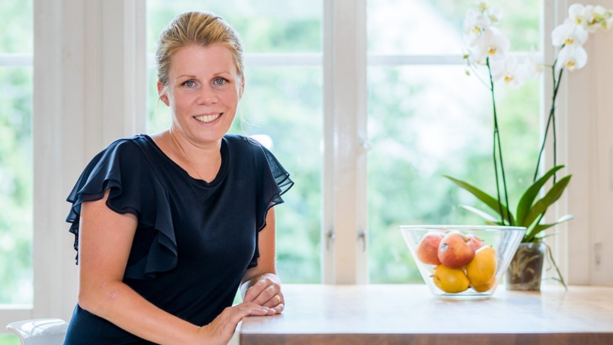 En kvinna i svart blus ler vid en köksbänk, med en skål med frukt och vita orkidéer i bakgrunden - kanske en stund tillsammans efter lektionerna på yrkeshögskolan.