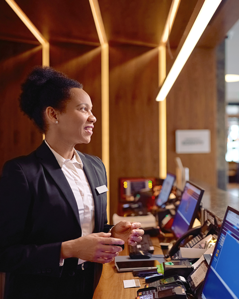 En hotellchef i kostym står vid receptionen och ler bredvid datorskärmarna och betalterminalerna.