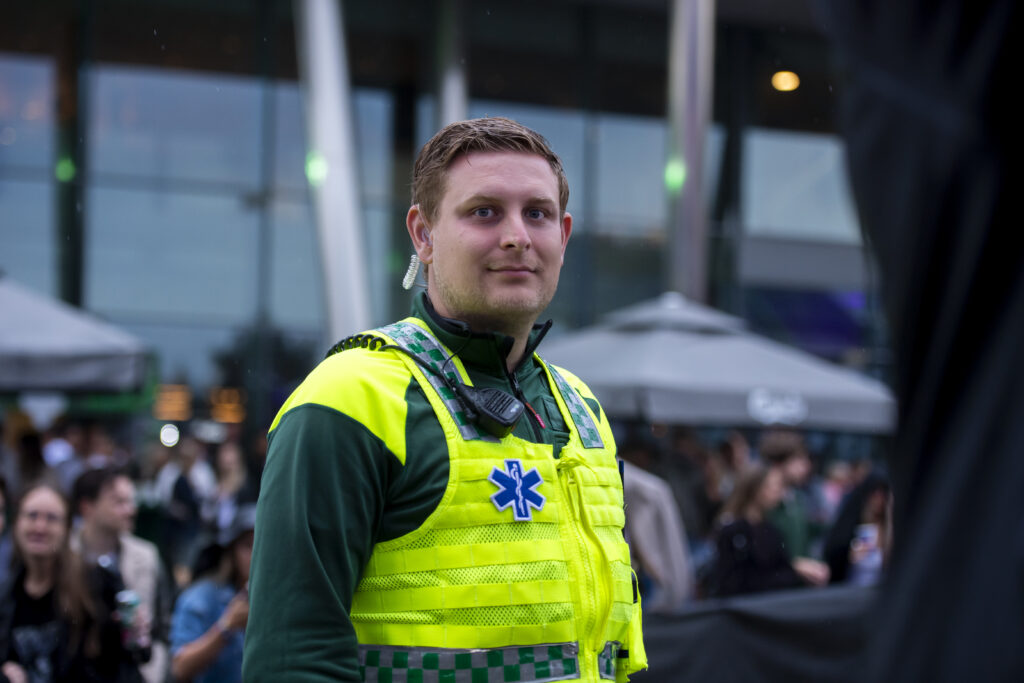 En sjukvårdare i gul väst står utanför ett event och representerar HLR-Tjänst och EventAmbulans AB i ledningsgruppen, med en folkmassa och tält i bakgrunden.