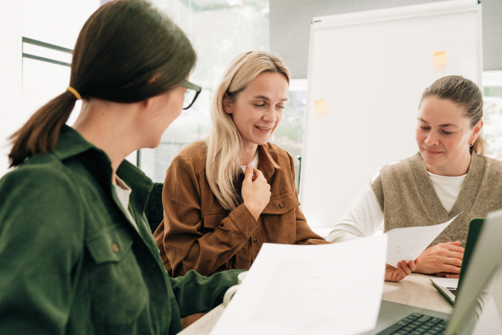 Tre kvinnor sitter vid ett bord och diskuterar papper, med en laptop och ett blädderblock i bakgrunden. YH Akademin störst på nationell yrkesutbildning - beviljas fem utbildningar, lyfts fram i samtalet.