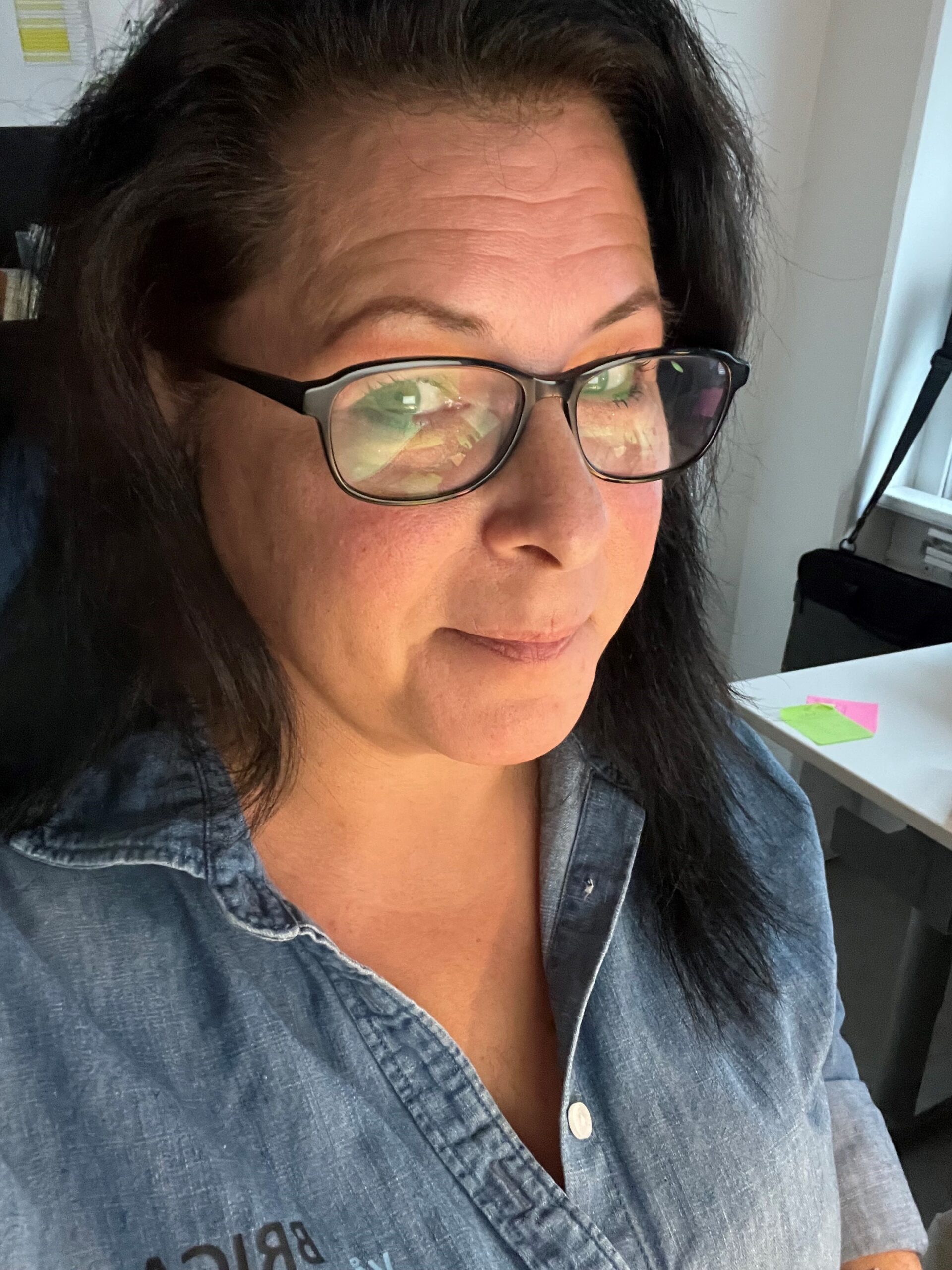 Kvinna med mörkt hår och glasögon, klädd i jeansskjorta, ler lite i ett starkt upplyst rum - kanske redo att gå med i Kurs: Hållbar affärsutveckling inom gårdsförsäljning.