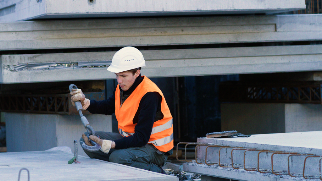 En byggnadsarbetare iförd skyddsväst och hjälm fäster en metallkrok i en betongplatta.