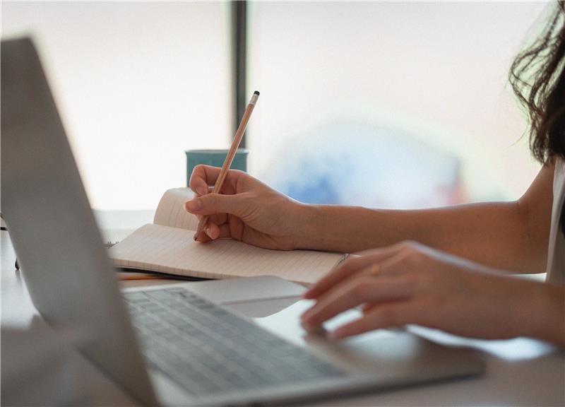 Person som skriver i en anteckningsbok bredvid en öppen laptop på ett skrivbord, med en kopp i bakgrunden - perfekt för studier eller planering av uppgifter på yrkeshögskola.