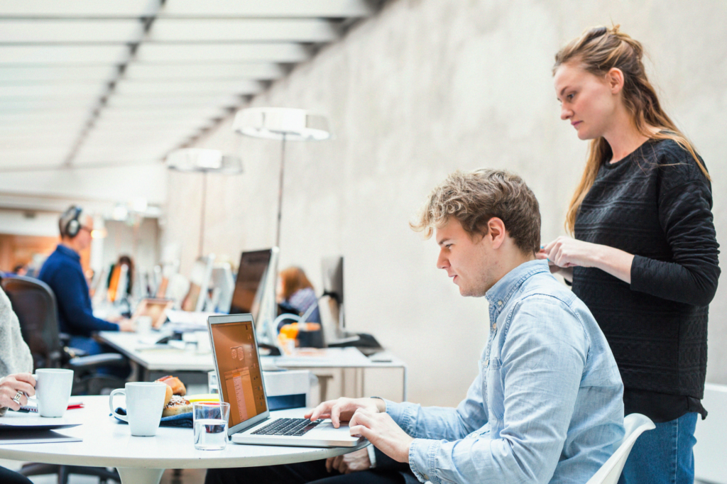 Två kollegor sitter vid en laptop i ett modernt, öppet kontor med andra som arbetar i bakgrunden och diskuterar studera utomlands - försäkring alternativ för ett kommande projekt.
