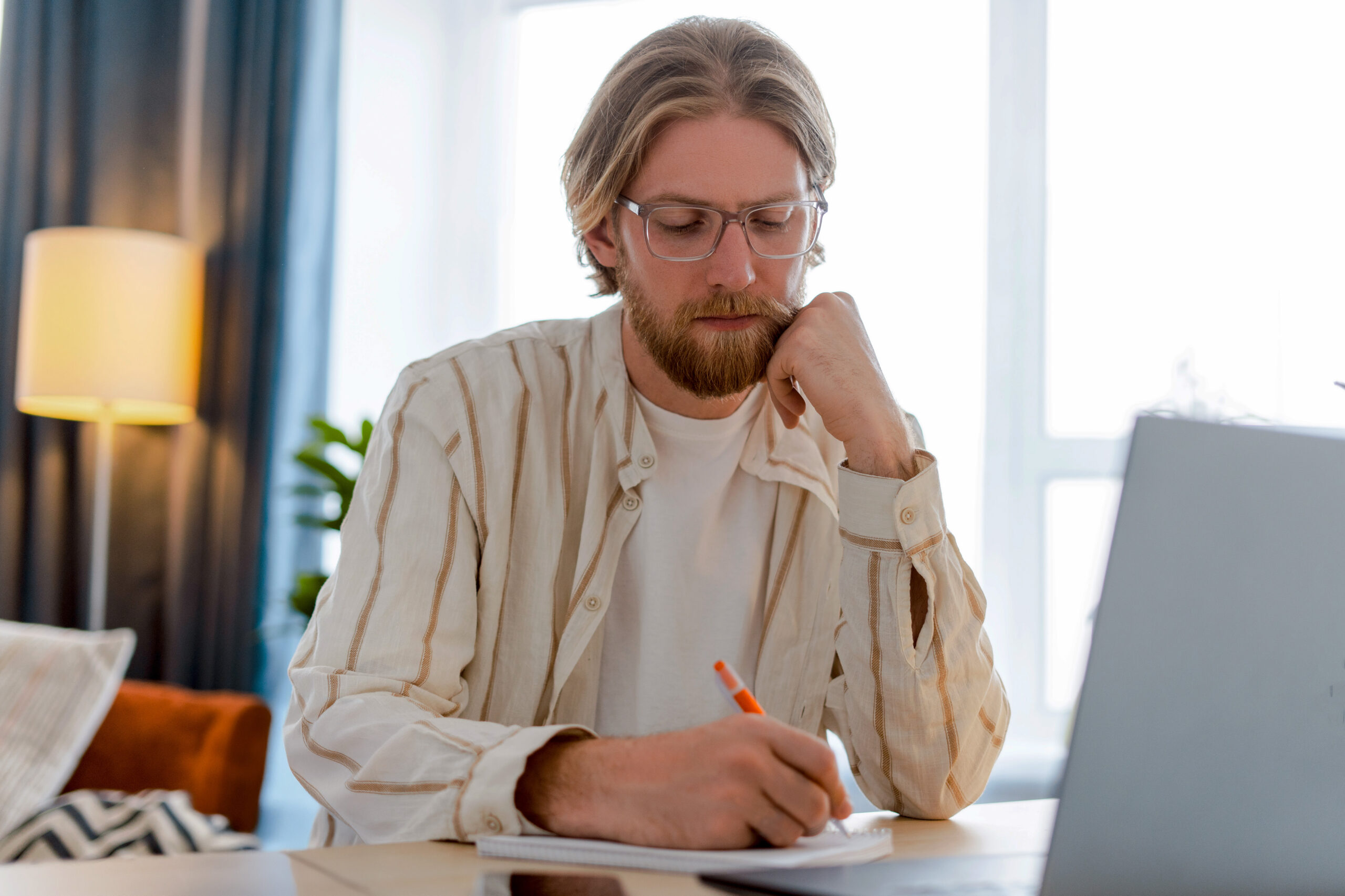 Man med glasögon skriver i en anteckningsbok medan han sitter vid ett skrivbord med en bärbar dator i ett ljust rum.