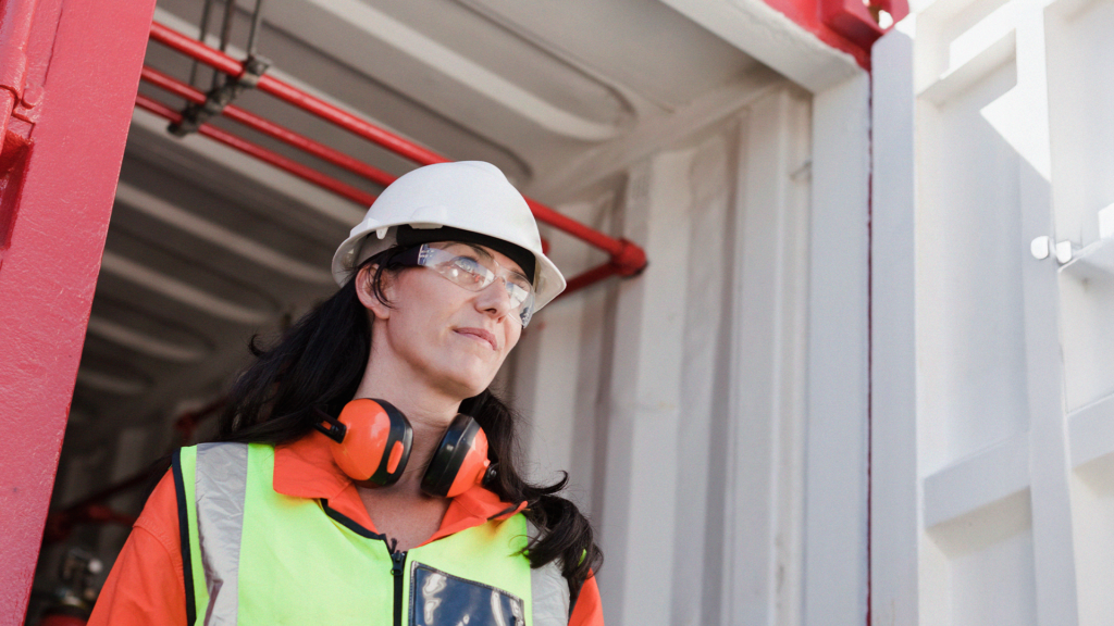 Kvinna i skyddsutrustning och skyddshjälm står inne i en industrimiljö och tittar åt sidan.