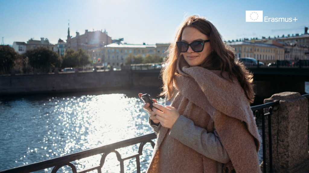 En kvinna i solglasögon och kappa står vid en flod och håller i sin telefon, kanske för att fånga minnen från sitt Erasmus+-äventyr, med stadens byggnader i bakgrunden.