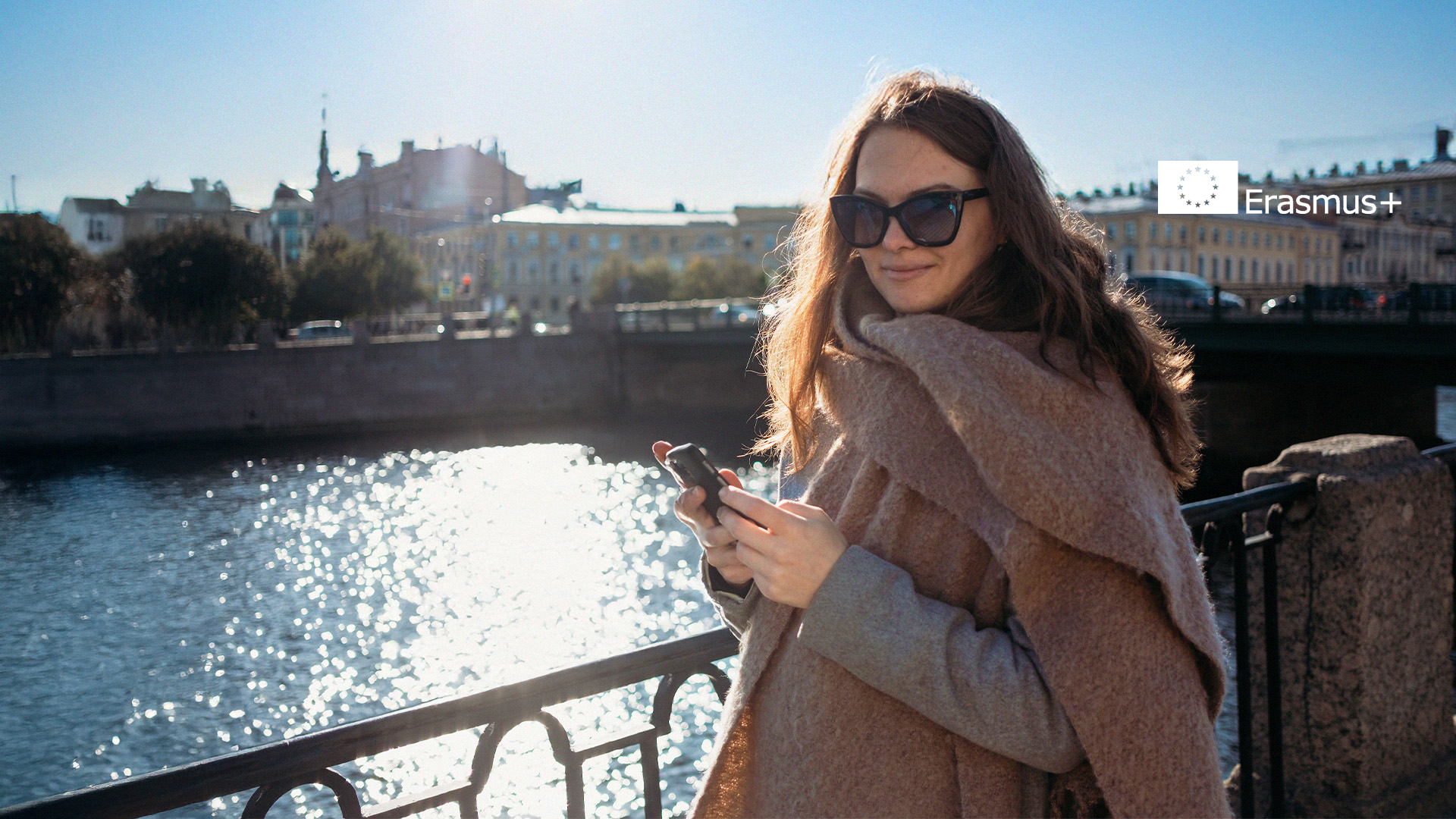 Kvinna med solglasögon vid en flod, med en telefon i handen och Erasmus+ logotypen i övre högra hörnet, som visar andan i Erasmus+ äventyr.