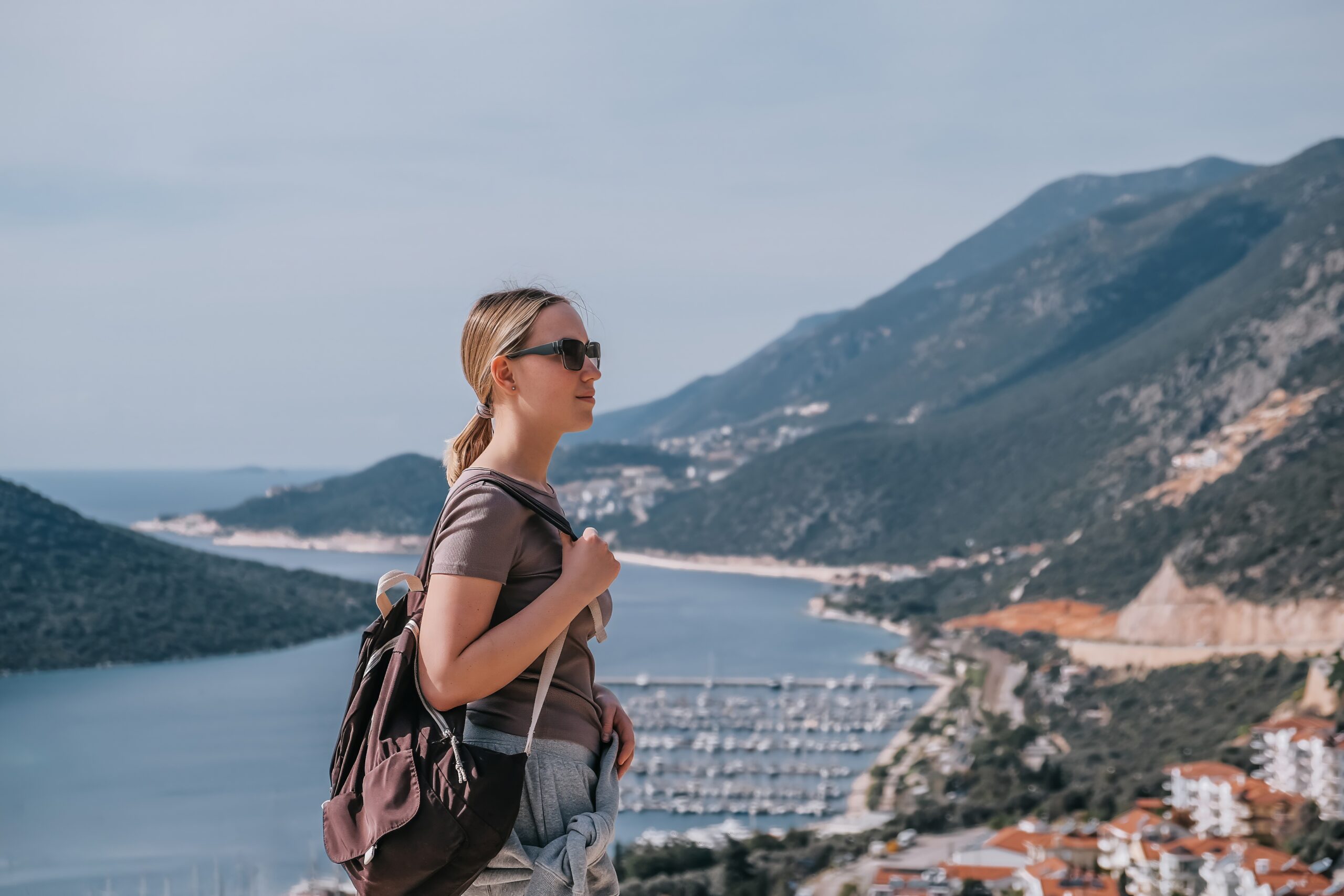 Kvinna med ryggsäck och solglasögon står med utsikt över en kuststad, marina och berg - perfekt inspiration för dig som planerar att studera utomlands - försäkring rekommenderas för sinnesfrid på ditt äventyr.