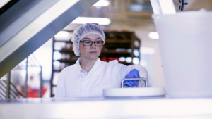 En laboratoriearbetare i vit rock, hårnät och handskar sköter utrustning i en ren, industriell miljö.