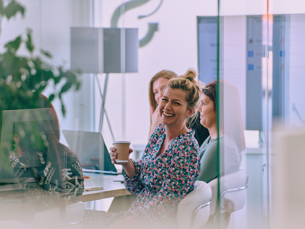 Kvinna ler och håller i en kaffekopp under ett möte med kollegor på ett modernt kontor.