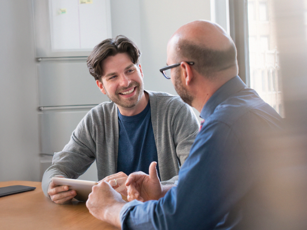 Två män sitter vid ett bord och har ett vänligt samtal; den ena håller i en surfplatta och ler.