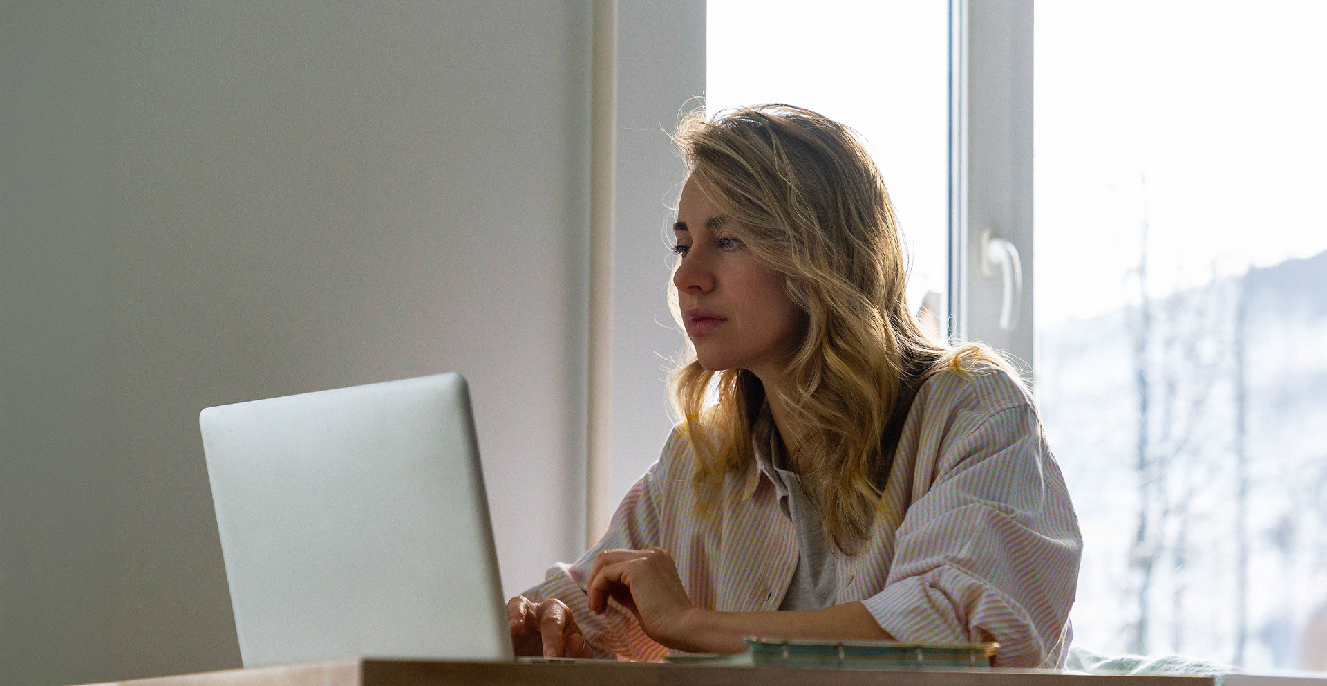 En kvinna sitter vid ett skrivbord med en bärbar dator nära ett fönster och naturligt ljus flödar in när hon studerar för sina yrkeshögskolekurser.