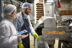 Två arbetare i skyddsutrustning inspekterar utrustning för livsmedelsbearbetning i en fabrik, den ena håller i en surfplatta.