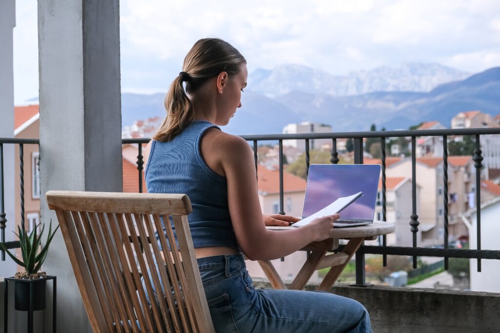 Kvinna arbetar på en laptop med en anteckningsbok på en balkong med utsikt över berg och hustak - perfekt för att planera studera utomlands och läsa om vikten av rätt försäkring.
