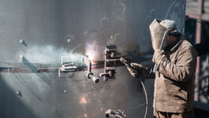 En arbetare i skyddsutrustning svetsar metall på en stor cylindrisk struktur, vilket ger upphov till gnistor och rök.