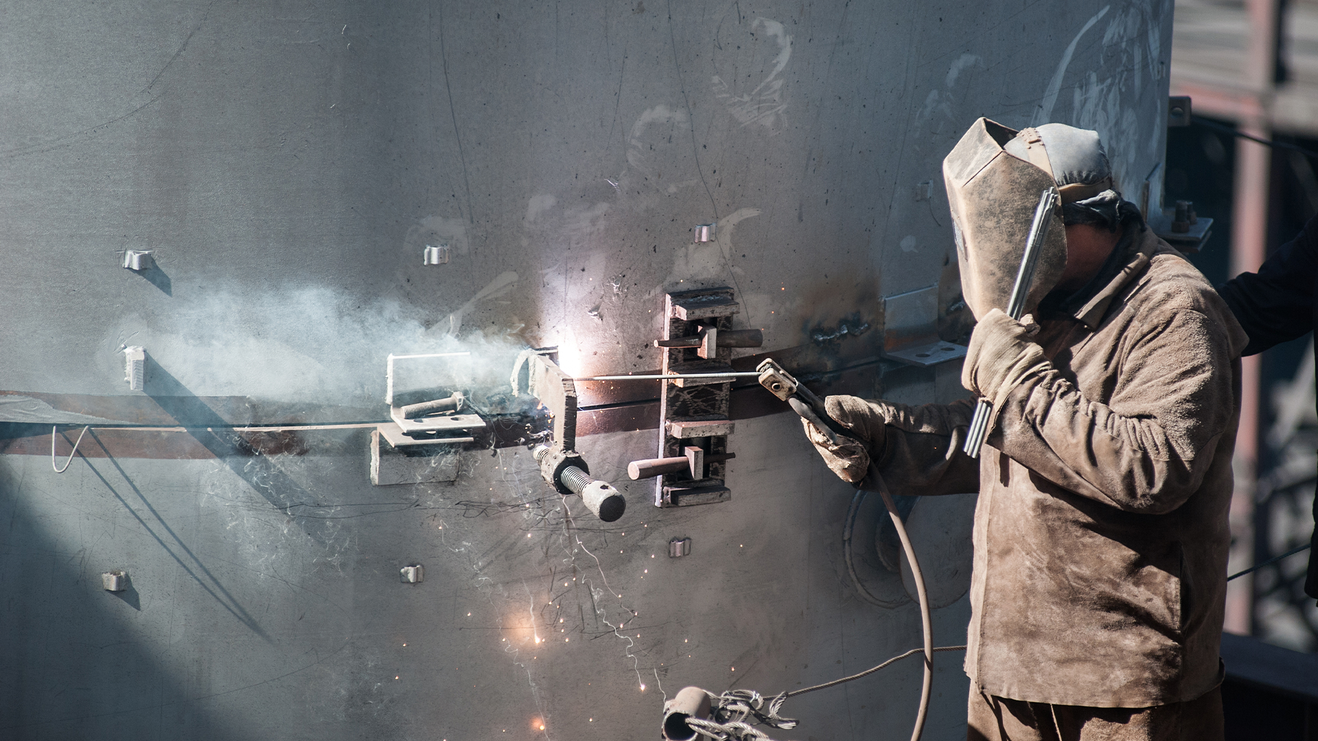 En arbetare i skyddsutrustning svetsar metall på en stor cylindrisk struktur, vilket ger upphov till gnistor och rök.