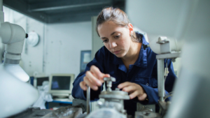 Kvinna i blå overall justerar maskiner i en industri- eller laboratoriemiljö.