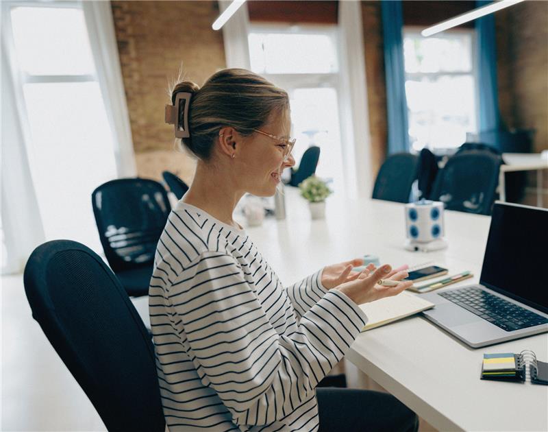Kvinna i randig skjorta sitter vid skrivbord och gestikulerar på sin laptop under ett videosamtal på en yrkeshögskola i ett ljust kontor.