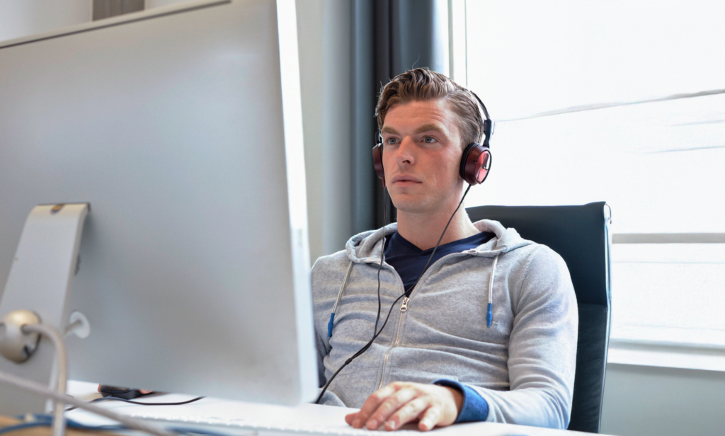 En man med hörlurar och luvtröja arbetar vid en dator i en ljus, modern kontorsmiljö, som förkroppsligar den fokuserade atmosfär som ofta finns på en yrkeshögskola.