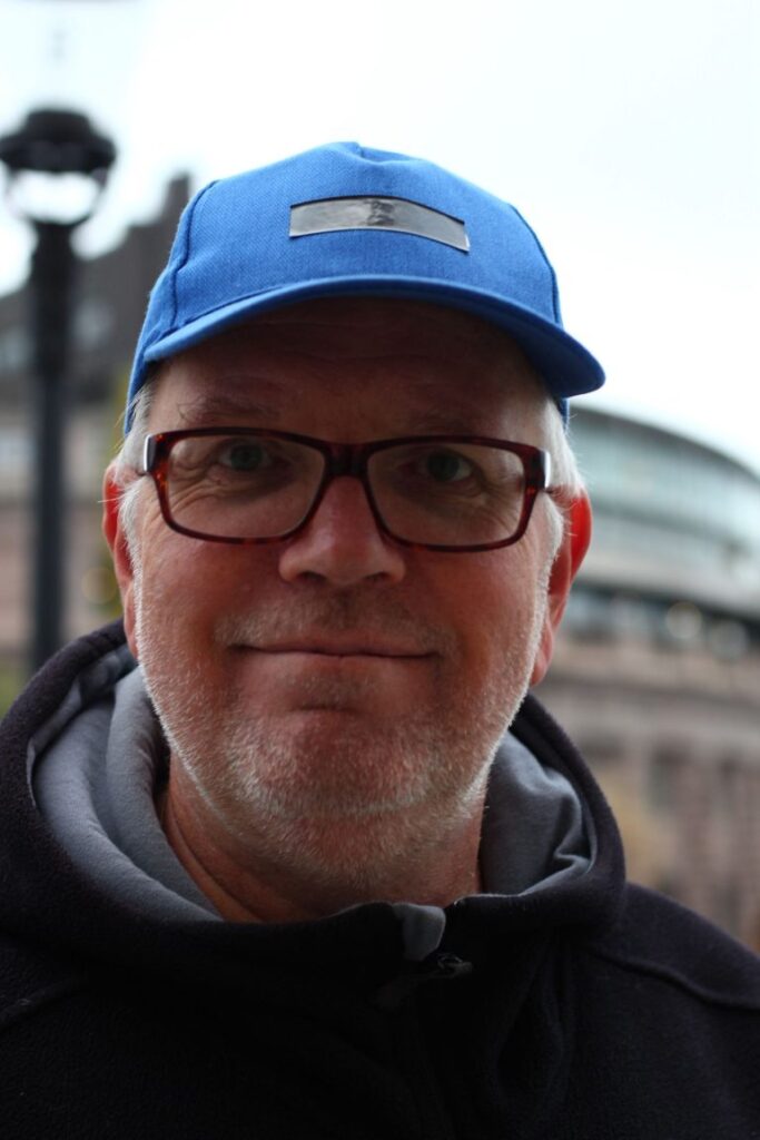 Man med glasögon och en blå keps ler utomhus; byggnader och en lyktstolpe i bakgrunden antyder hans professionella atmosfär som Customer Success Manager, perfekt för din LinkedIn i ledningsgruppen-profil.