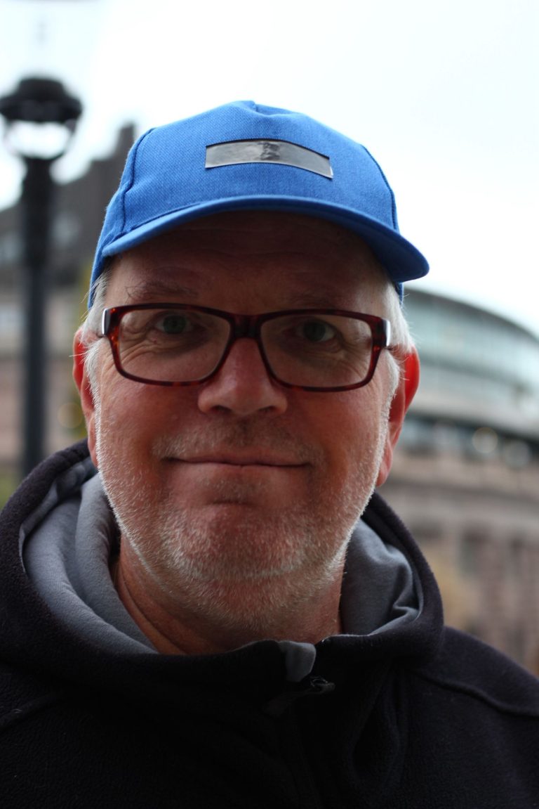 Man med glasögon och en blå keps ler utomhus; byggnader och en lyktstolpe i bakgrunden antyder hans professionella atmosfär som Customer Success Manager, perfekt för din LinkedIn i ledningsgruppen-profil.