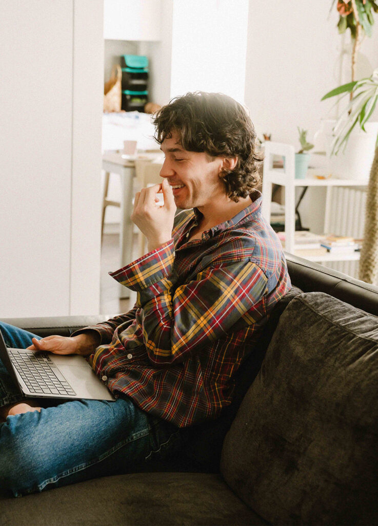 En leende person i rutig skjorta sitter i en soffa med en bärbar dator i ett ljust, mysigt vardagsrum och surfar på kurser från YH Akademin.