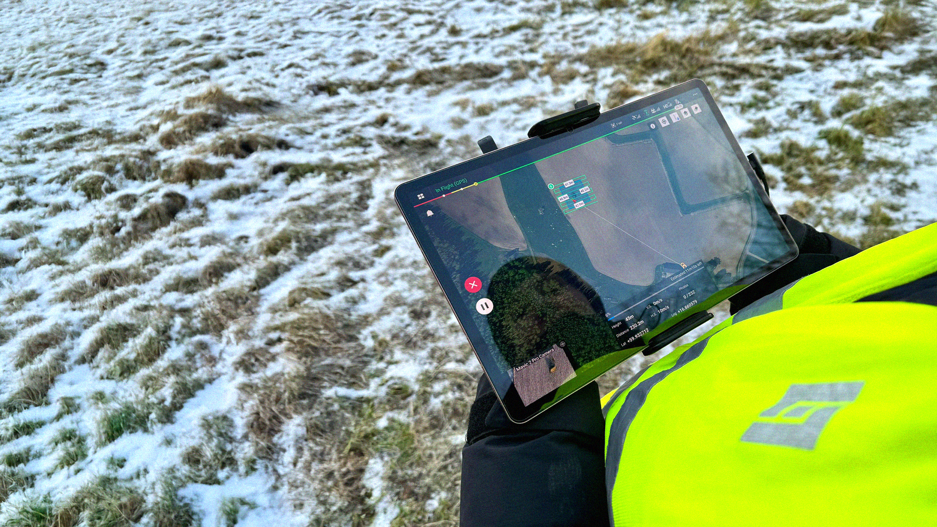 En person i gul väst styr en drönare med hjälp av en surfplatta över ett snöigt gräsfält.