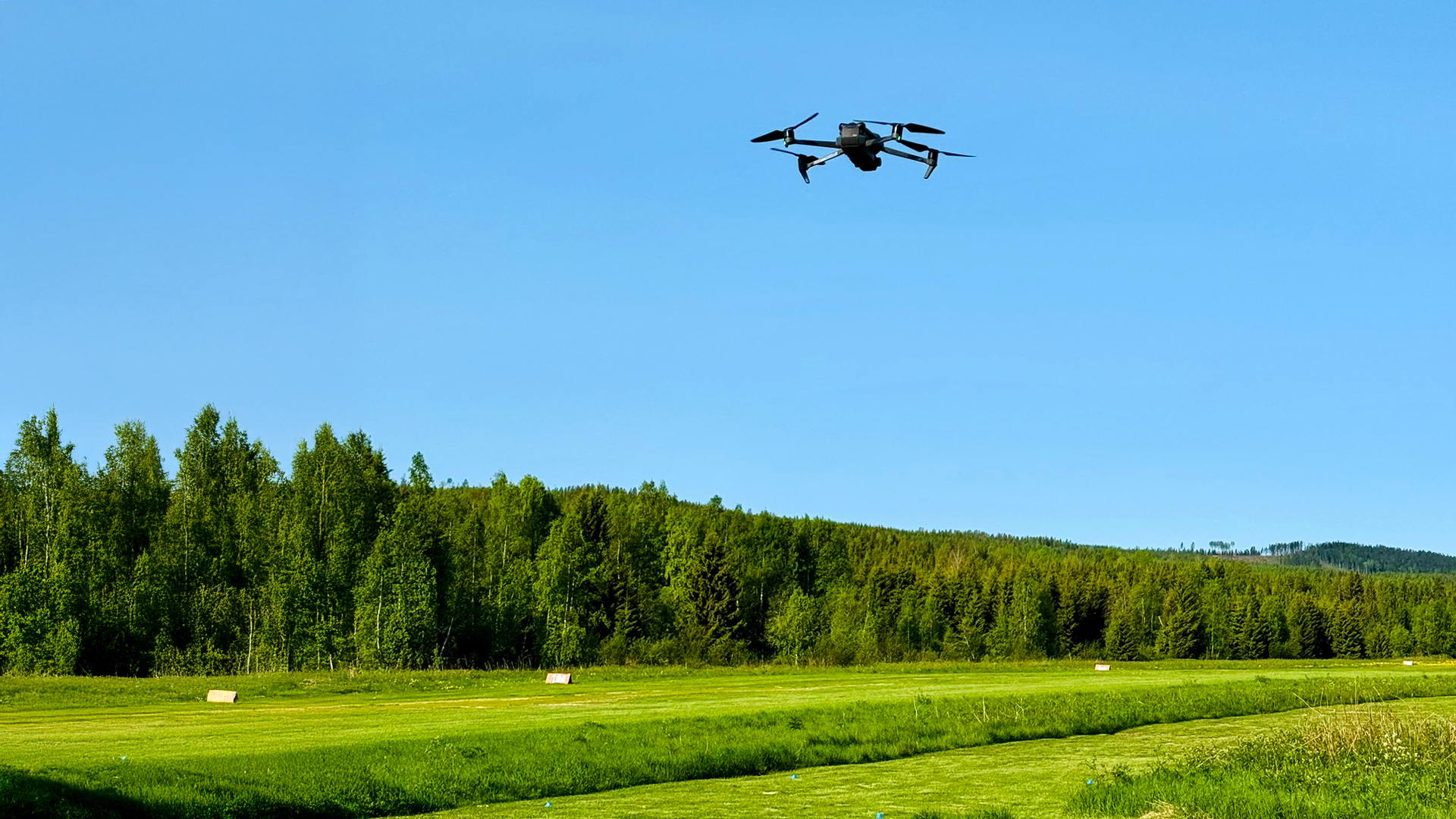 En svart drönare flyger över ett gräsbevuxet fält med träd och en klarblå himmel i bakgrunden.