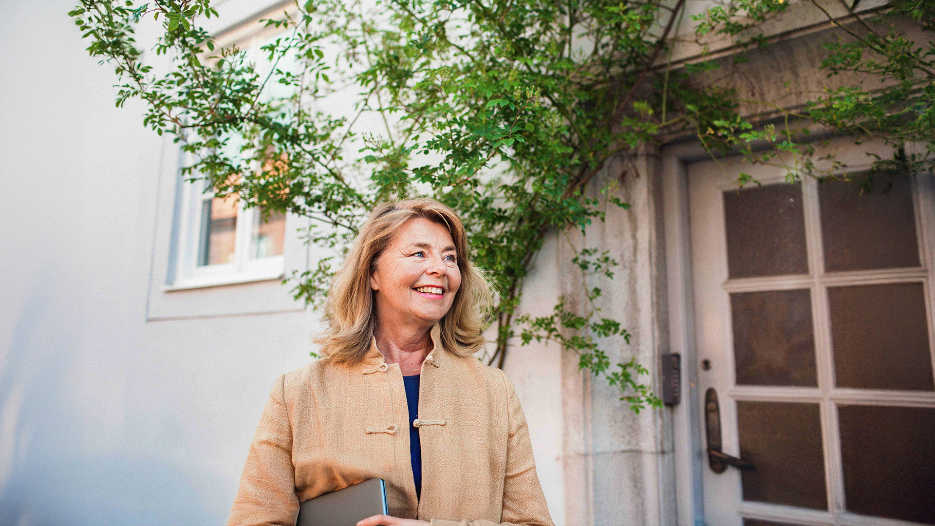 En leende kvinna i beige jacka står utomhus med en bärbar dator i handen nära en byggnad med gröna vinrankor - kanske på väg till en kurs på YH Akademin.