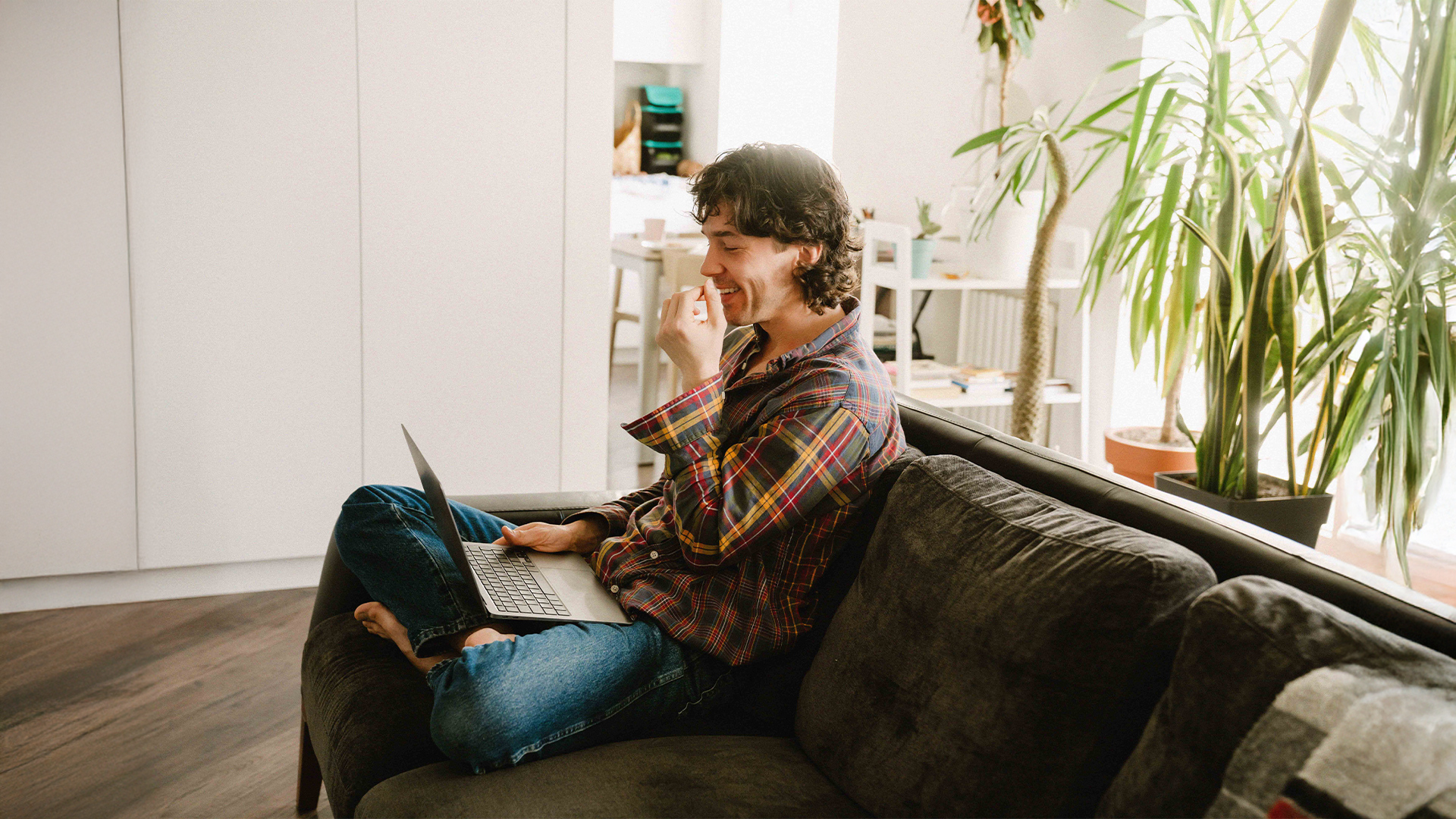 Person som sitter i en soffa med en laptop och ler, omgiven av krukväxter i ett ljust rum - utforskar kurser från YH Akademin.