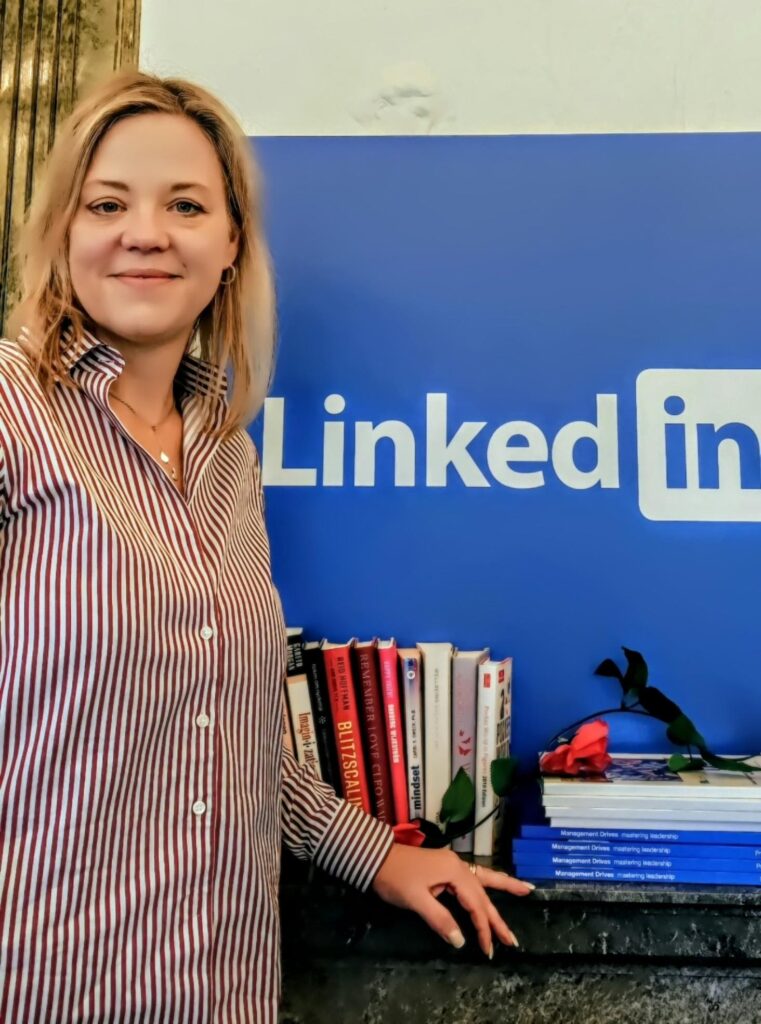Kvinna står vid en LinkedIn-skylt, med böcker och en enda röd ros på disken bredvid sig. Hon representerar HLR-Tjänst och EventAmbulans AB i ledningsgruppen.