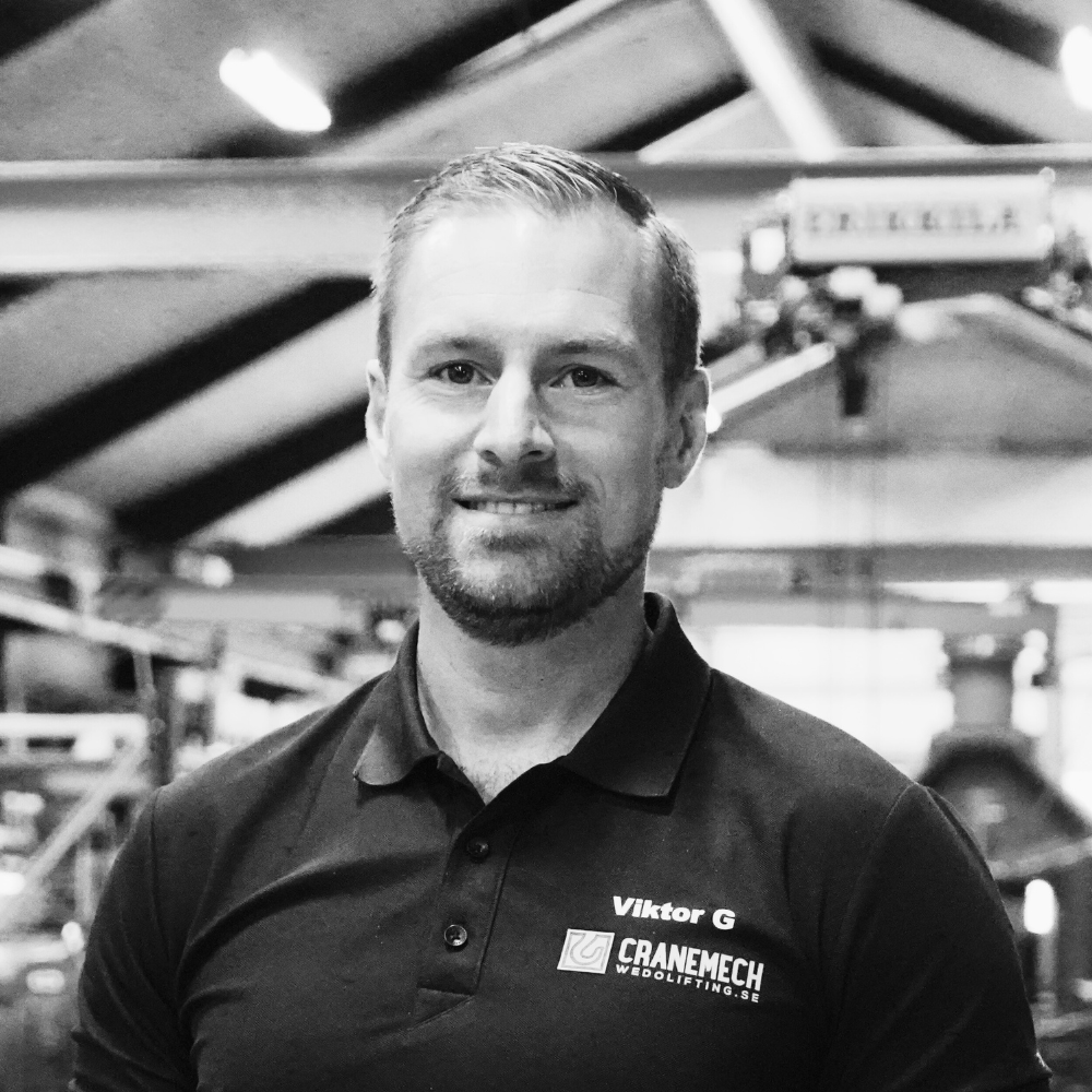 Man i svart polotröja som står och ler i en industriell verkstadsmiljö och förkroppsligar professionalismen hos en Kran- och traverstekniker.