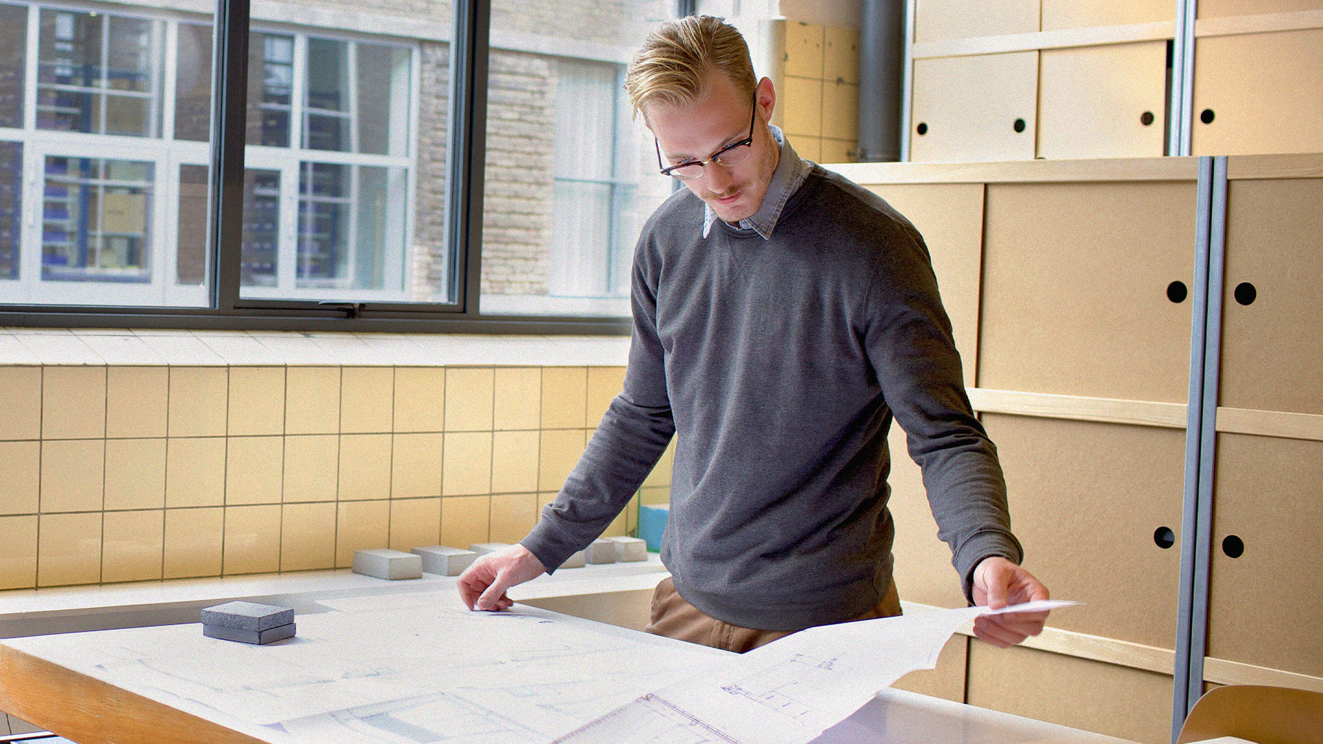 En person granskar arkitektritningar på ett skrivbord i en modern, väl upplyst kontorslokal och fokuserar på brandskydd för byggprojektledare och platschefer för att säkerställa att säkerheten uppfylls.