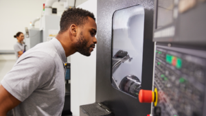 En man i grå skjorta tittar på en CNC-maskin som är i drift; en annan person, möjligen en CNC-tekniker med utbildning inom additiv tillverkning (YH-flex), arbetar i bakgrunden i liknande klädsel.