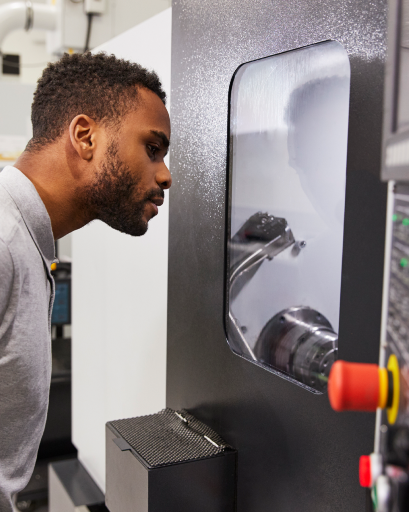 En CNC-tekniker med inriktning på additiv tillverkning inom YH-flex står och tittar genom ett fönster mot maskinerna inne i en industri- eller tillverkningsanläggning.