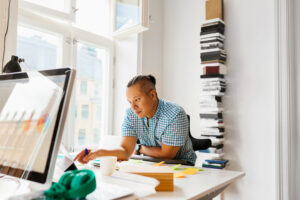 En person i rutig skjorta arbetar vid ett skrivbord fyllt med papper, klisterlappar och en hög trave böcker - försjunken i uppgifter inom teknisk projektledning.