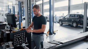 En man i svart skjorta använder en dator i en bilverkstad, omgiven av bilar, när han arbetar med fordonselektronikdiagnostik.