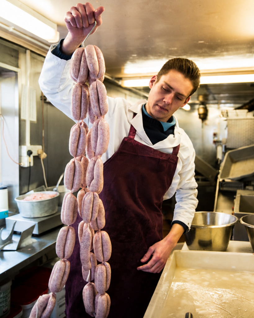 En student på utbildningen ”Köttförädlare YH-flex” i förkläde håller upp en lång rad råa korvar i ett kök.