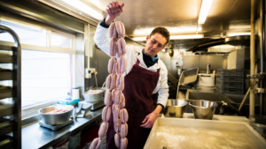 En man i förkläde håller upp en lång rad råa korvar i ett storkök och visar upp sina färdigheter som en äkta Köttförädlare YH-flex.