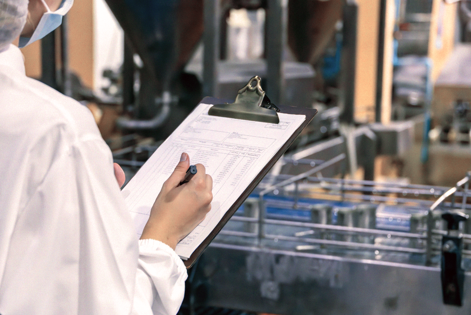 Person i labbrock med skrivplatta och penna inspekterar utrustning i en industri- eller laboratoriemiljö och visar den noggranna kvalitetsmedvetenhet som förväntas av en köttförädlare.