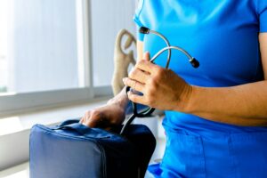 En person i blå operationskläder håller ett stetoskop och en sjukvårdsväska nära ett fönster, redo att ge omvårdnad vid diabetes.